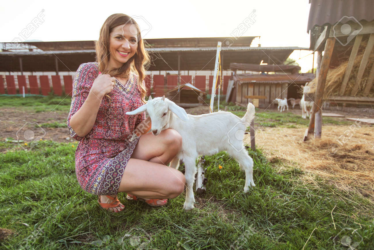 168512891-young-woman-crouching-playing-with-goat-kid-smiling-farm-behind-her-.jpg